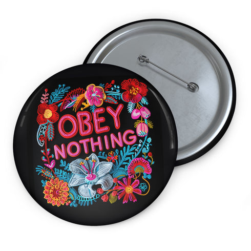 A black round pinback button featuring floral embroidery-style designs and bold red text that reads "Obey Nothing."